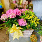 Floral arrangement with pink and yellow flowers in a basket, decorated with a yellow bow, in a garden setting.
