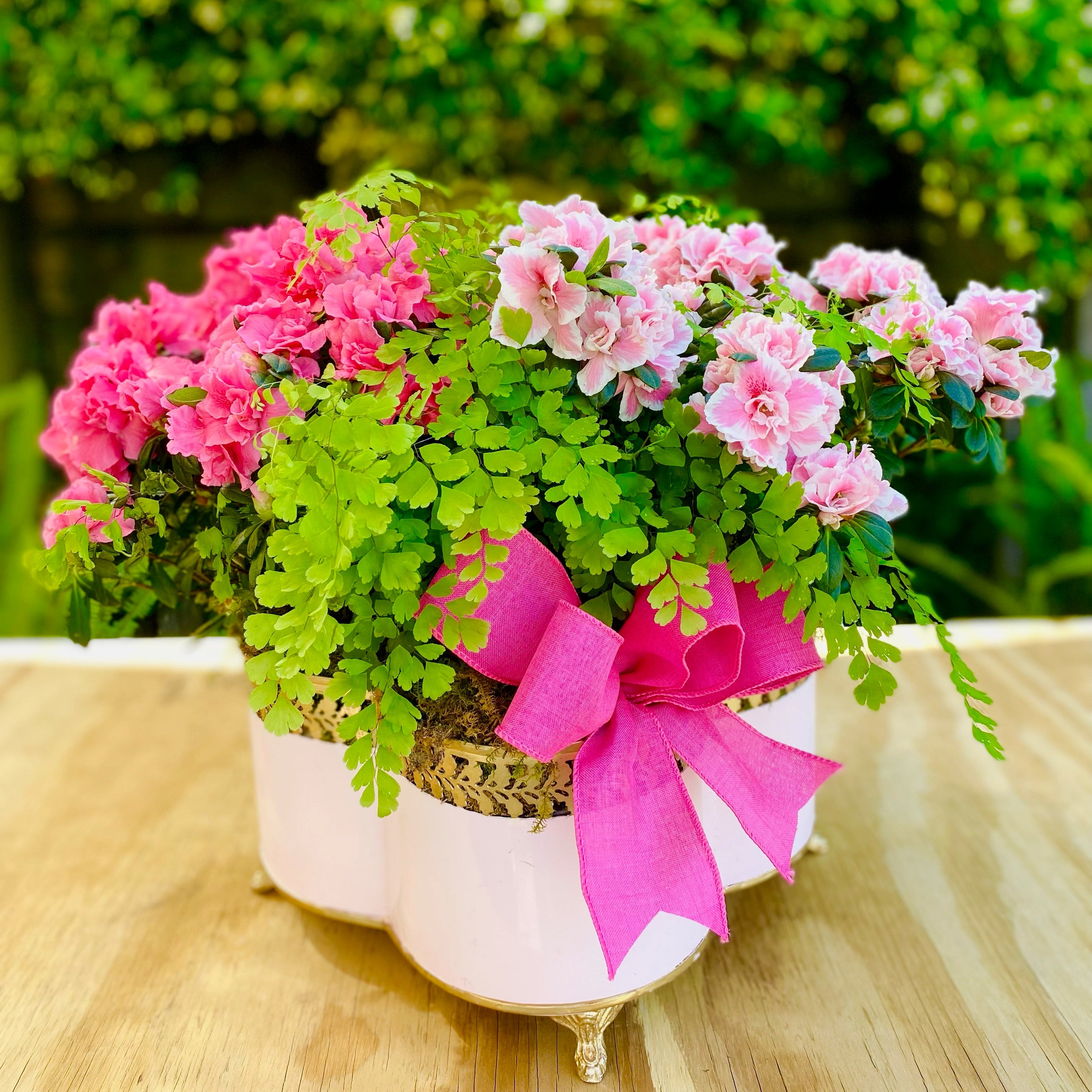 Pink container planted with pink flowers and greenery, tied with a pink ribbon on a wooden surface.