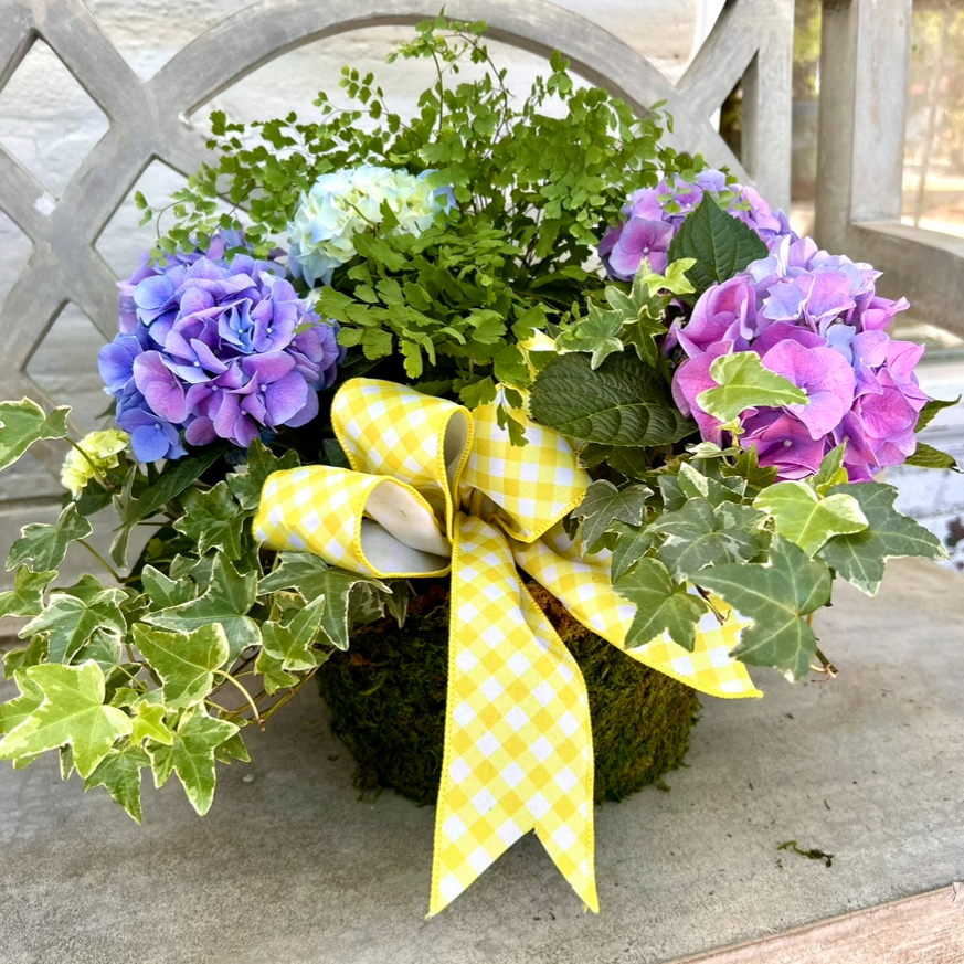 Floral arrangement with purple and blue flowers and a yellow bow on a concrete bench.